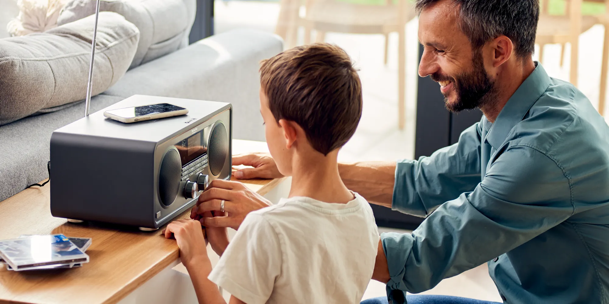 Ein Kind und sein Vater knien fröhlich vor einem DAB+ Radio im Wohnzimmer. Ein gemeinsamer Hörspaß in digitaler Qualität.