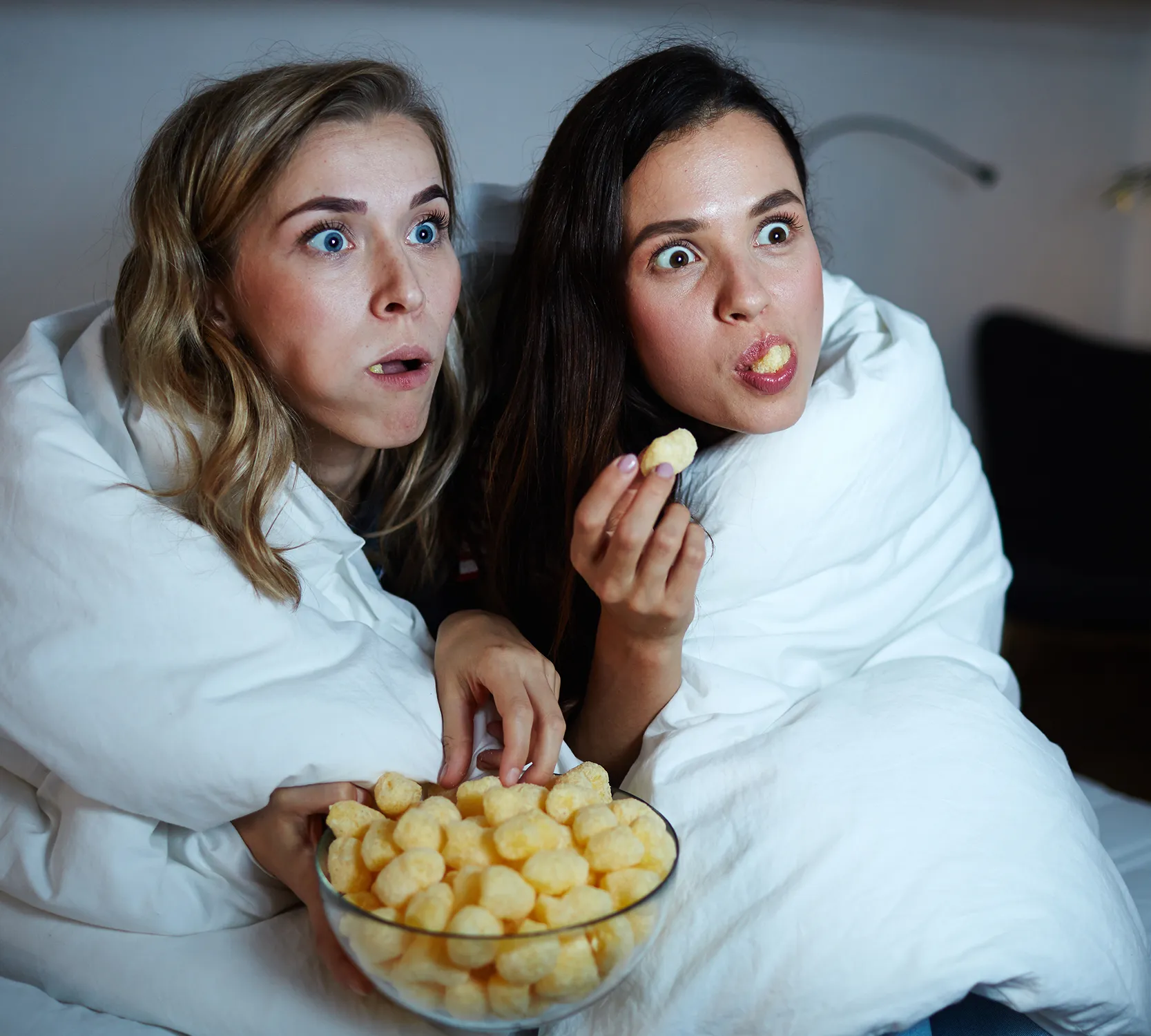 Image of two women sitting excitedly with wide eyes in front of a TV, wrapped in a blanket and eating.